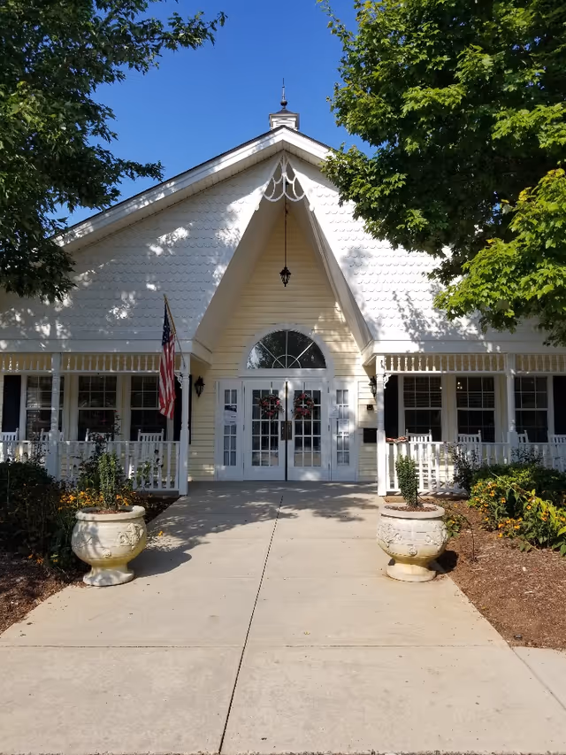 Front entrance of a white, cottage-style building with a peaked roof, double glass doors, porch, American flag and two large planters.