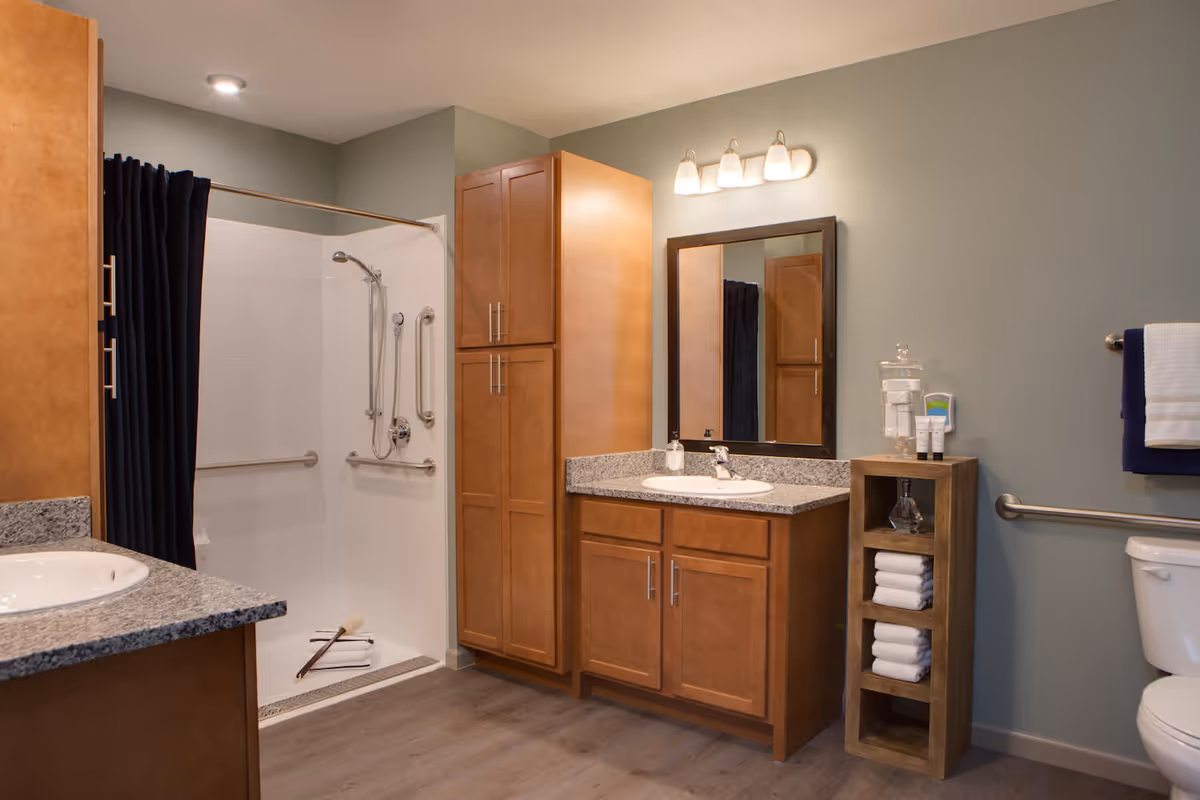 A clean and modern bathroom featuring a walk-in shower with grab bars and a dark shower curtain, a wooden vanity with a granite countertop and sink, a large mirror with three light fixtures above it, a tall wooden cabinet, a small wooden shelf holding neatly folded white towels and toiletries, and a toilet with a grab bar beside it. The walls are painted a soft green color and the floor has a wood-like finish.