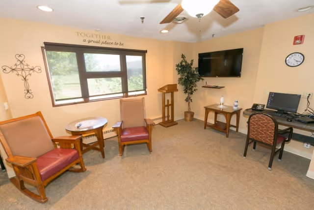 A cozy room with two wooden armchairs with red cushions, a small round wooden table between them, a window with a view outside, a wall-mounted TV, a small wooden table with a lamp and a water bottle, a computer desk with a chair and a desktop computer, a potted plant, a ceiling fan with lights, and a wall clock. The wall above the window has the phrase 'TOGETHER a great place to be'.