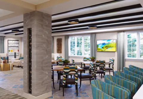 A spacious, well-lit common area with multiple tables and chairs featuring colorful geometric patterns. Large windows with curtains allow natural light to fill the room. A flat-screen TV is mounted on the wall displaying a scenic landscape. The room has a modern design with a decorative column and ceiling lights.