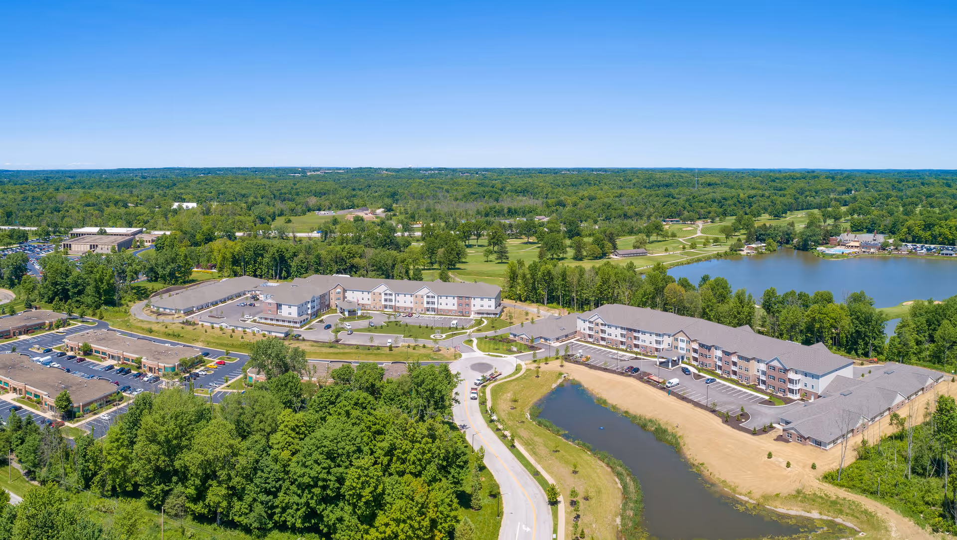 Aerial view of the Danbury Hudson senior living facility surrounded by greenery, a pond, and parking areas under a clear blue sky.