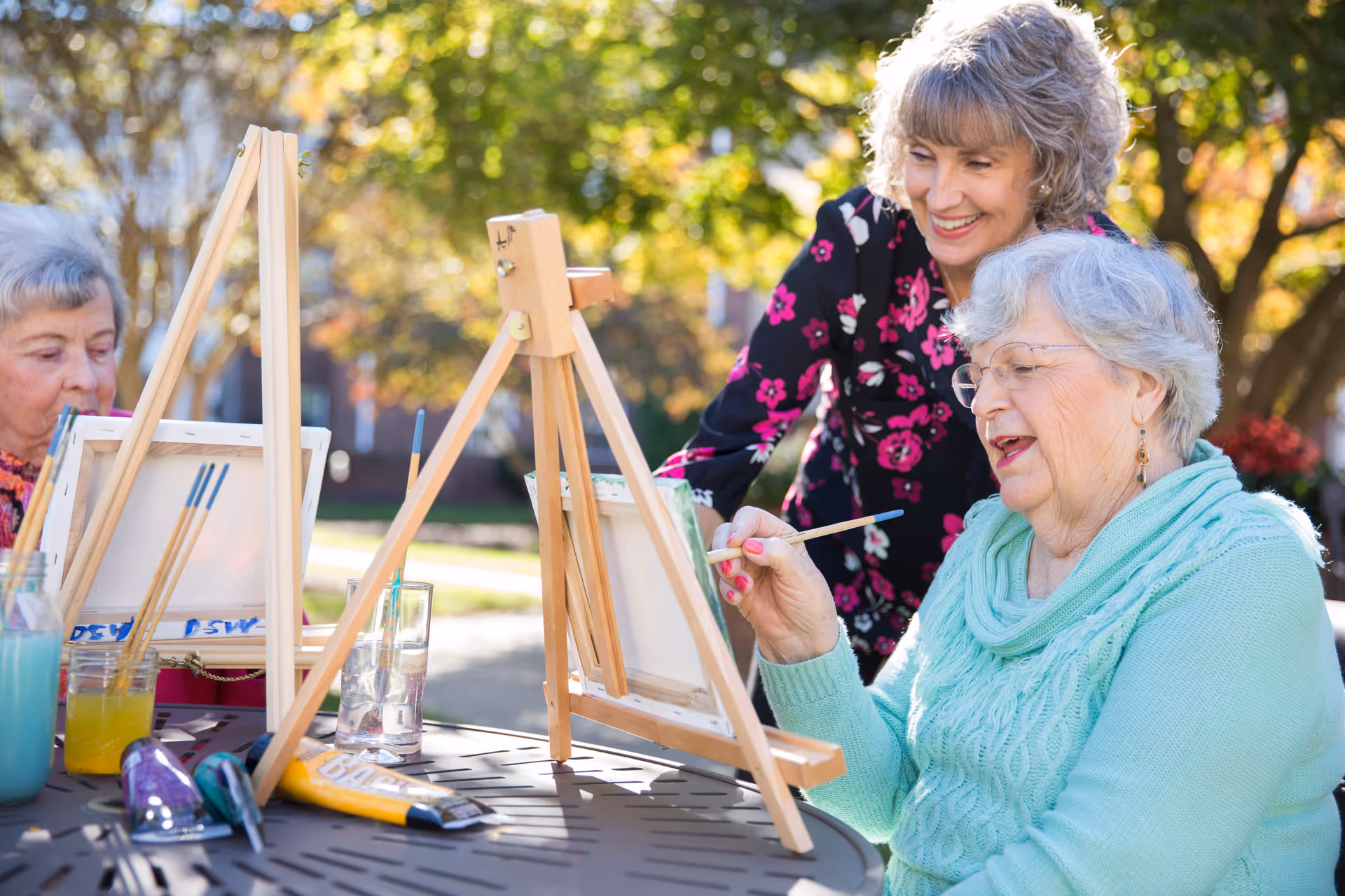 Three elderly women outdoors at a table engaged in painting on canvases set on easels. One woman in a turquoise sweater is holding a paintbrush and smiling, while another woman in a floral top looks on supportively. Art supplies including paint tubes and brushes are on the table, with trees and sunlight in the background.