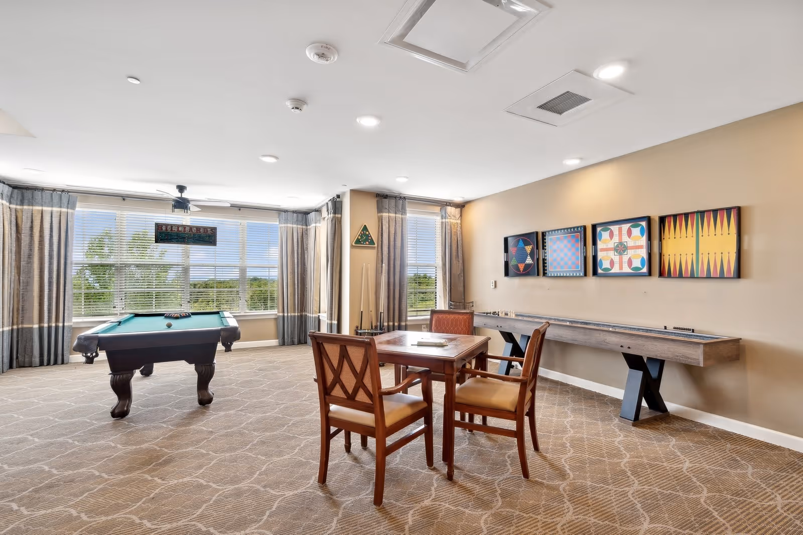 A spacious game room with a pool table, a shuffleboard table, and a card table with four chairs. The room has large windows with curtains allowing natural light to fill the space. Colorful game-themed artwork is displayed on the wall above the shuffleboard table.