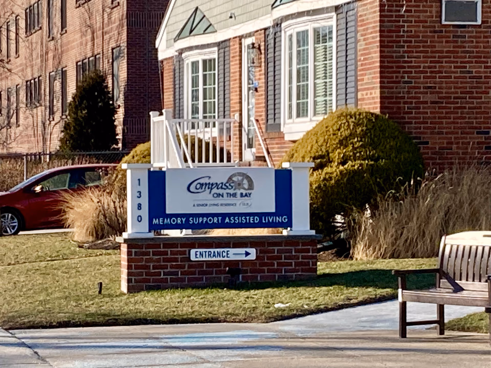 Outdoor view of a brick building with a sign in front that reads 'Compass on the Bay Memory Support Assisted Living' with the address number 1380 and an entrance arrow. There is a wooden bench on the right side and some bushes and grass around the sign.