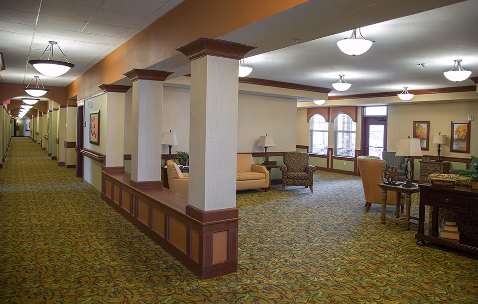 Carpeted senior living facility common area with lounge seating, decorative columns, and a long hallway.