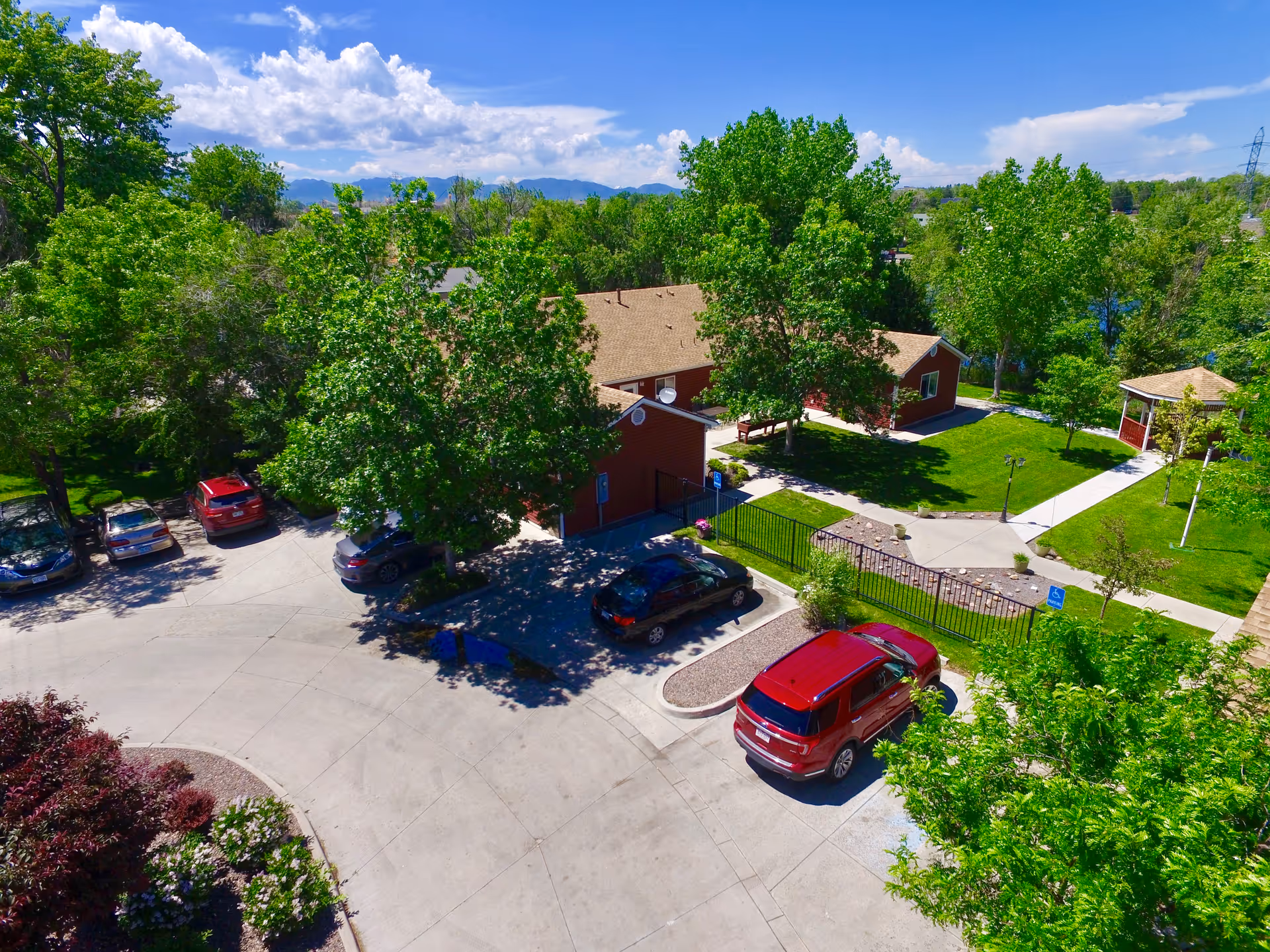 Aerial view of a tree-lined assisted living complex with red single-story buildings, parked cars and landscaped lawns and walkways.
