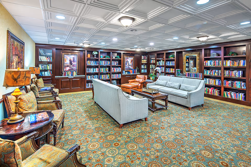 A cozy library room with wooden bookshelves filled with books lining the walls. The room features two light gray sofas facing each other with a wooden coffee table in between, a brown leather armchair with an ottoman, and several upholstered chairs along the left wall. The carpet has a detailed floral pattern, and the ceiling has a white coffered design with recessed lighting. There are framed paintings and decorative plants on the shelves and walls.