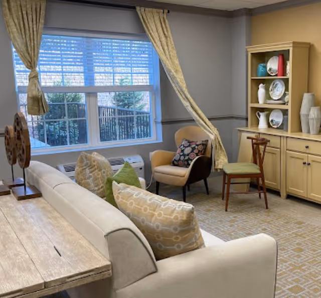 A cozy living room area with a beige sofa adorned with patterned pillows, a wooden table behind the sofa, a beige armchair with a floral cushion, a wooden chair with a green cushion, and a beige cabinet displaying decorative plates and vases. A large window with beige curtains lets in natural light, showing some greenery outside.