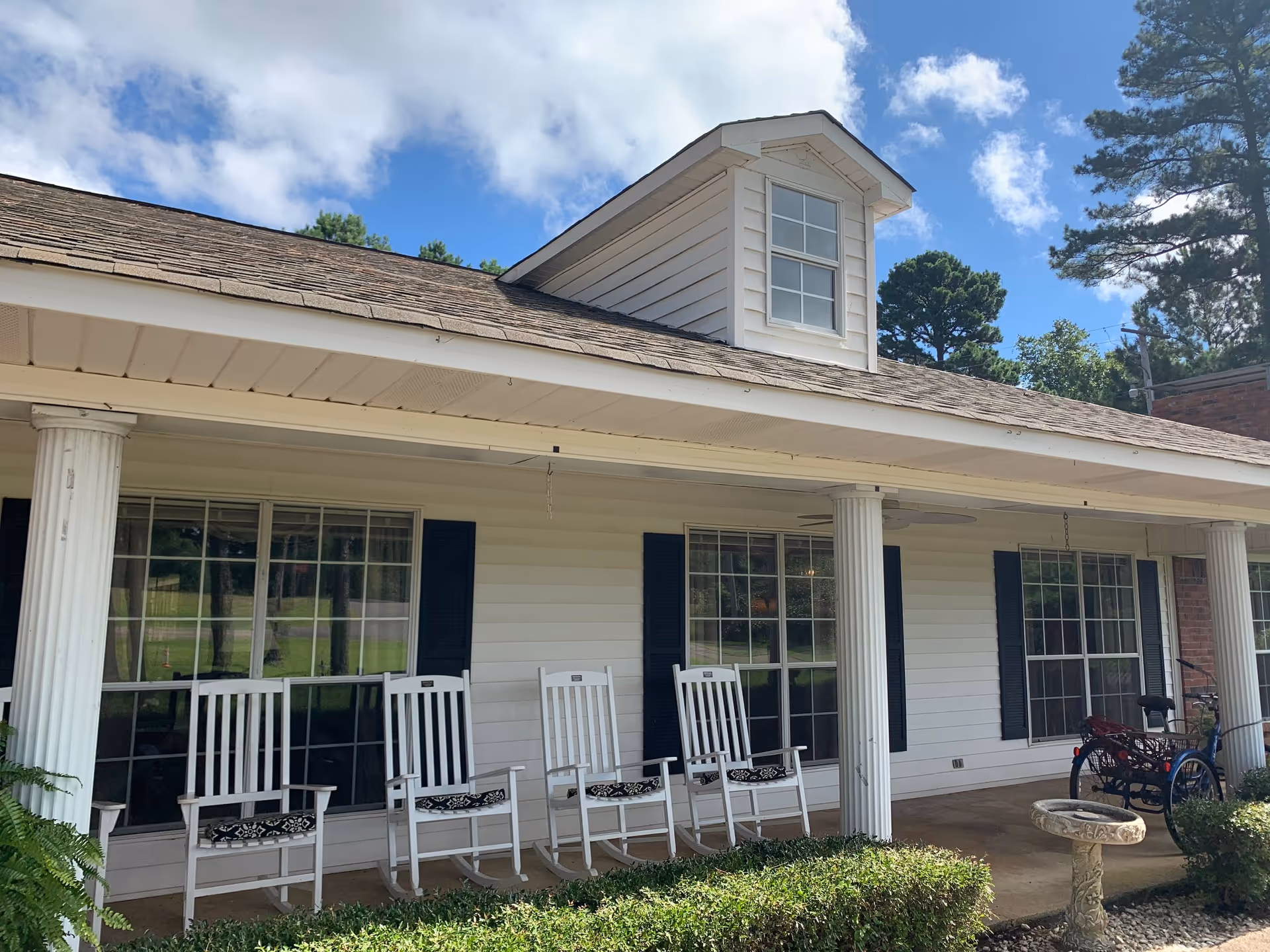 Front porch of a white building with four white rocking chairs with cushions, large windows with black shutters, white columns supporting the roof, a birdbath in the garden, and a tricycle parked on the right side. The sky is partly cloudy with blue patches.