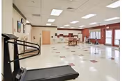 A spacious rehabilitation and nursing center room with a treadmill in the foreground, tables and chairs in the middle, and a kitchenette area in the background. The room has a tiled floor with a pattern of red and white squares, fluorescent ceiling lights, and large windows letting in natural light.