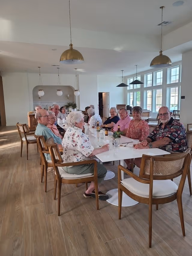 A group of senior residents seated around a long dining table in a bright communal dining room.