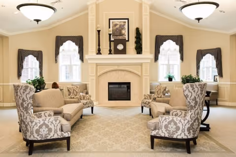 A spacious and elegant living room with beige walls and carpet. The room features a central fireplace with decorative items on the mantel, including candles, a clock, and a framed picture. There are four patterned armchairs and two beige sofas arranged symmetrically around the fireplace. Three windows with white curtains allow natural light to enter the room. The ceiling has two large light fixtures.