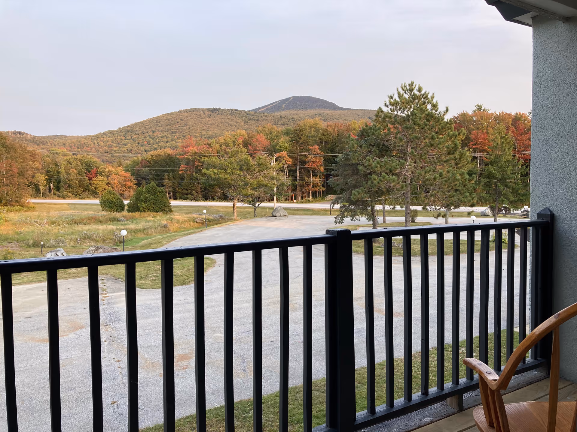 View from a balcony with black railing overlooking a parking area, trees with autumn foliage, and a mountain in the background under a clear sky.