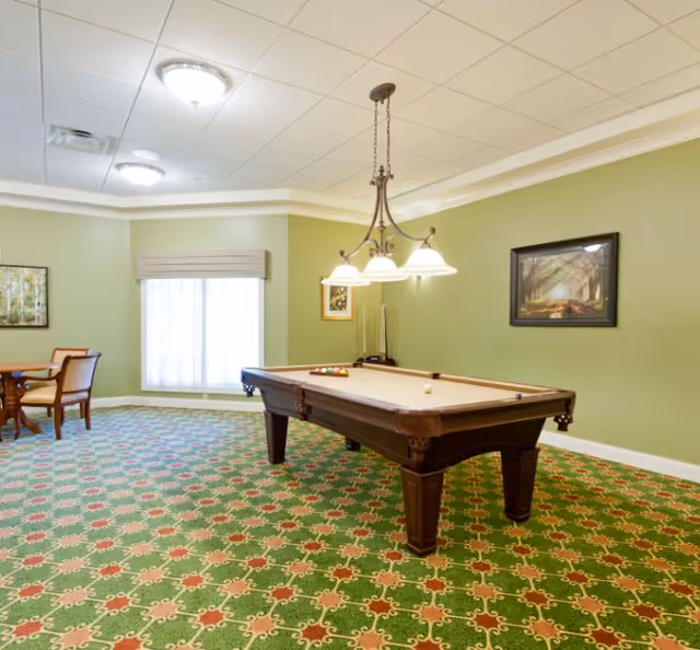 A recreational room with a pool table in the center, overhead lighting fixture above it, green patterned carpet, light green walls with framed artwork, and a small table with chairs near a window with a beige valance.