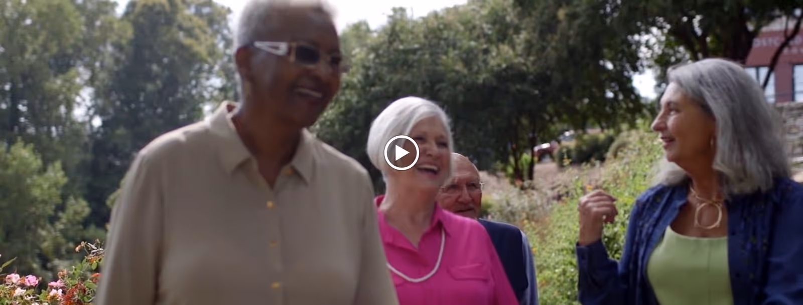 A group of four older adults walking outdoors along a garden path, smiling and engaging in conversation with trees and greenery in the background.