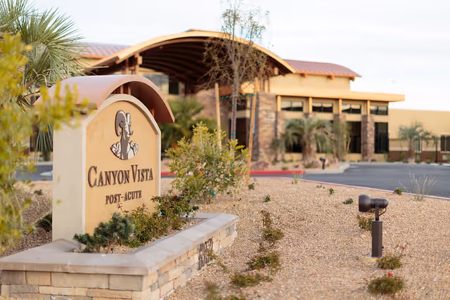 Outdoor view of the Canyon Vista Post-Acute facility entrance with a stone sign displaying the facility name and a landscaped area with desert plants and gravel in the foreground. The building features a curved roof and large windows.