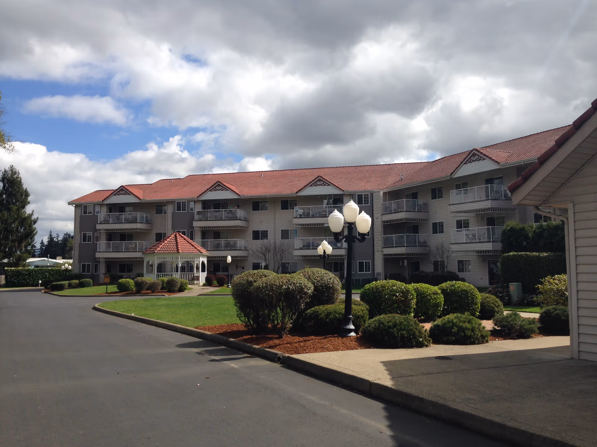 Exterior view of a multi-story senior living facility with balconies, a red-tiled roof, a gazebo in the courtyard, landscaped bushes, and lamp posts under a partly cloudy sky.