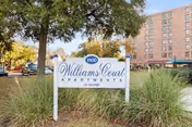Outdoor view of the Thomas F Williams Court Apartments sign surrounded by grass and trees, with a multi-story apartment building and parked cars visible in the background.
