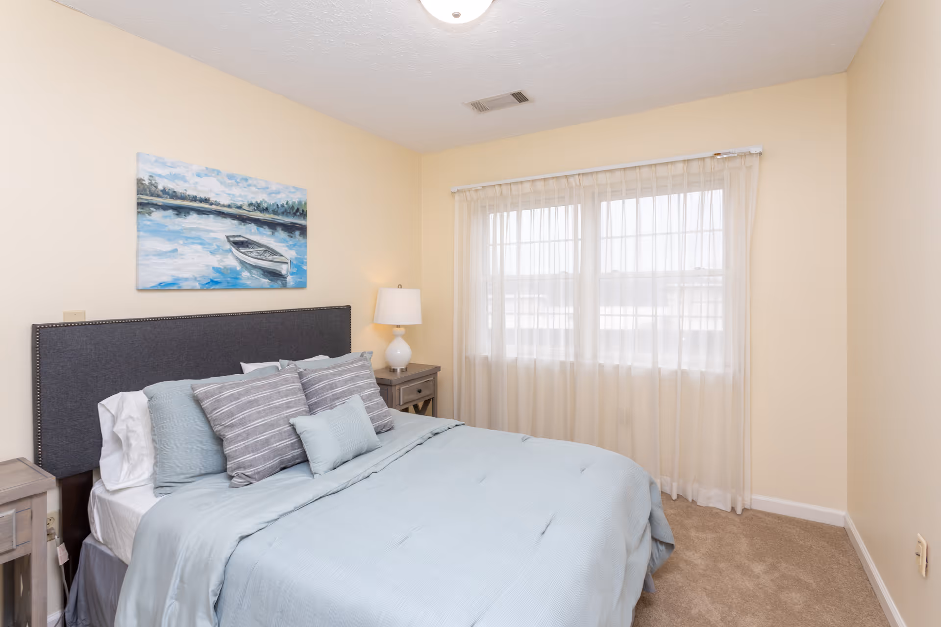 A bedroom with a neatly made bed featuring light blue bedding and multiple pillows. There is a dark gray upholstered headboard, two wooden nightstands with a white lamp on the right side, a large window with sheer white curtains, and a painting of a boat on water hanging above the bed.
