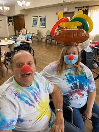 Two residents in tie-dye shirts wearing red clown noses—one with inflatable rings on her head—sit in a communal dining/activity room with other residents in the background.