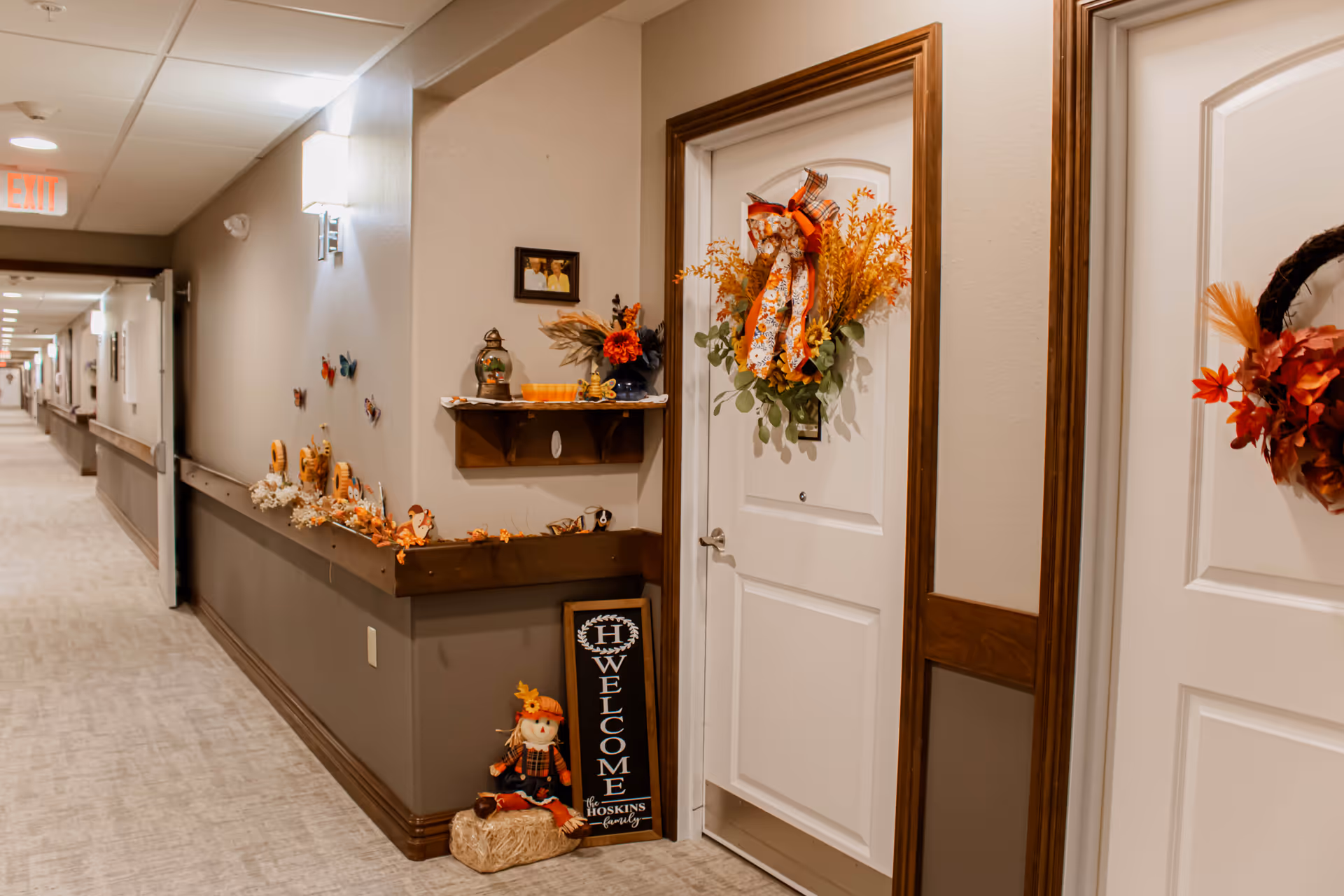 A decorated hallway in an assisted living facility with white doors adorned with autumn-themed wreaths. The hallway has beige walls with a wooden chair rail and a light carpet. Seasonal decorations including a scarecrow, a welcome sign, and small fall-themed ornaments are placed along a wooden ledge and on the floor near the doors. The hallway extends into the distance with an exit sign visible at the far end.