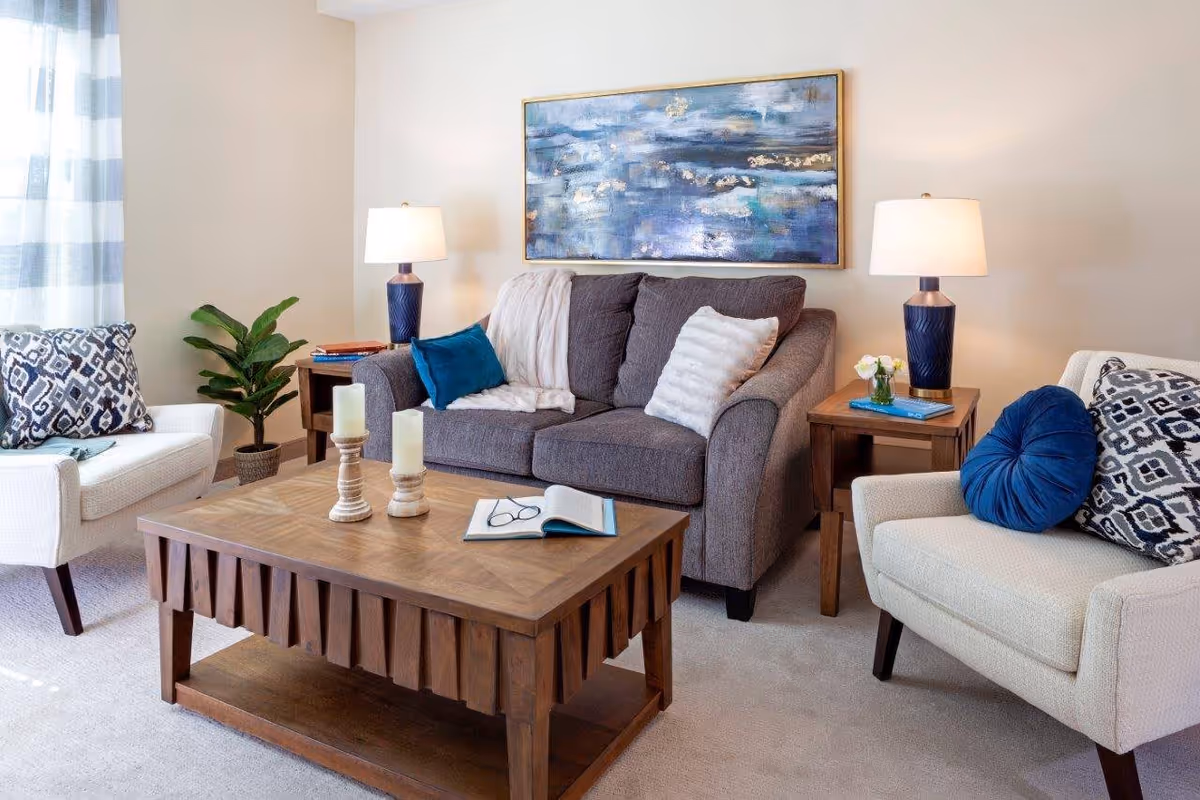 A cozy living room with a gray sofa adorned with white and blue pillows, two beige armchairs with patterned and solid blue cushions, a wooden coffee table with candles and an open book, two wooden side tables each with a lamp and decorative items, a potted plant, and a large abstract painting on the wall.