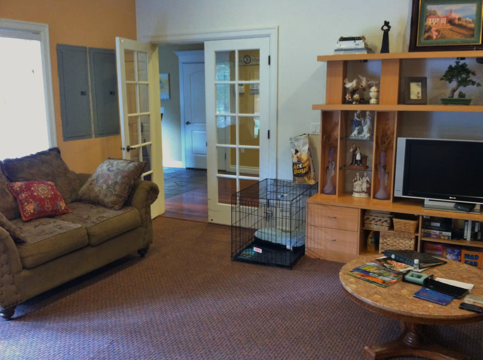 A cozy living room with a patterned sofa adorned with decorative pillows, a wooden entertainment center with shelves displaying figurines and a small TV, a round coffee table with magazines and other items, and a pet crate on the carpeted floor near glass-paneled double doors leading to another room.