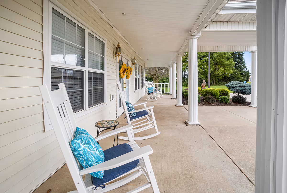 Covered porch area with white rocking chairs featuring blue cushions and patterned pillows, small round side tables, beige siding walls, white pillars, and decorative wreaths on the doors. Green shrubs and trees are visible in the background.