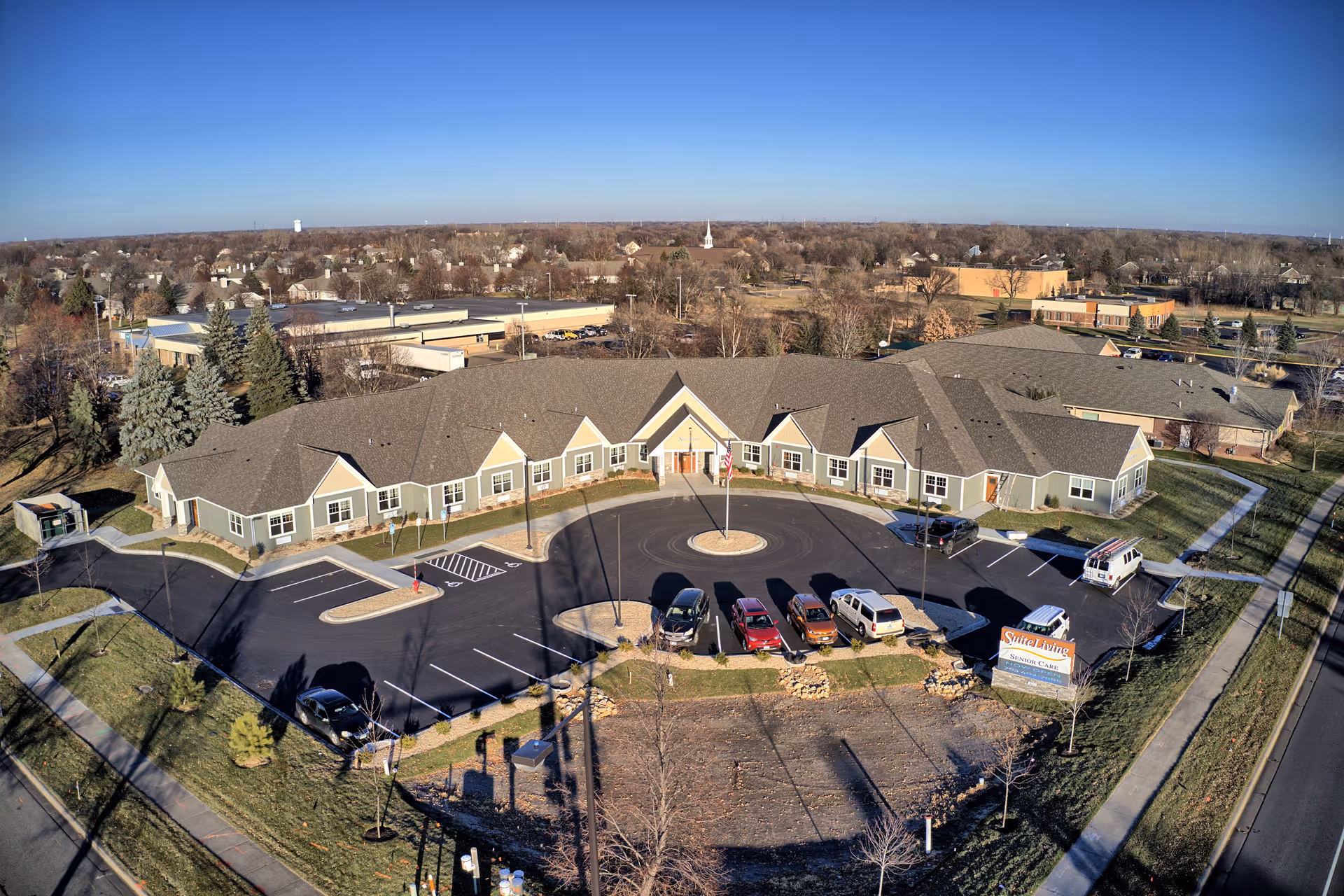 Aerial view of Suite Living Senior Care of West St. Paul building with a circular driveway and parking lot in front. The building is single-story with a gray roof and light green exterior walls. Several cars are parked in the lot, and there is a sign near the entrance. Surrounding the building are trees, sidewalks, and a suburban neighborhood under a clear blue sky.