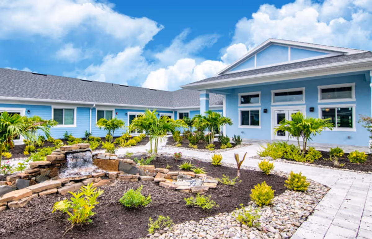 Exterior view of a light blue assisted living facility building with a landscaped garden featuring small palm trees, shrubs, a stone water fountain, and paved walkways under a partly cloudy blue sky.