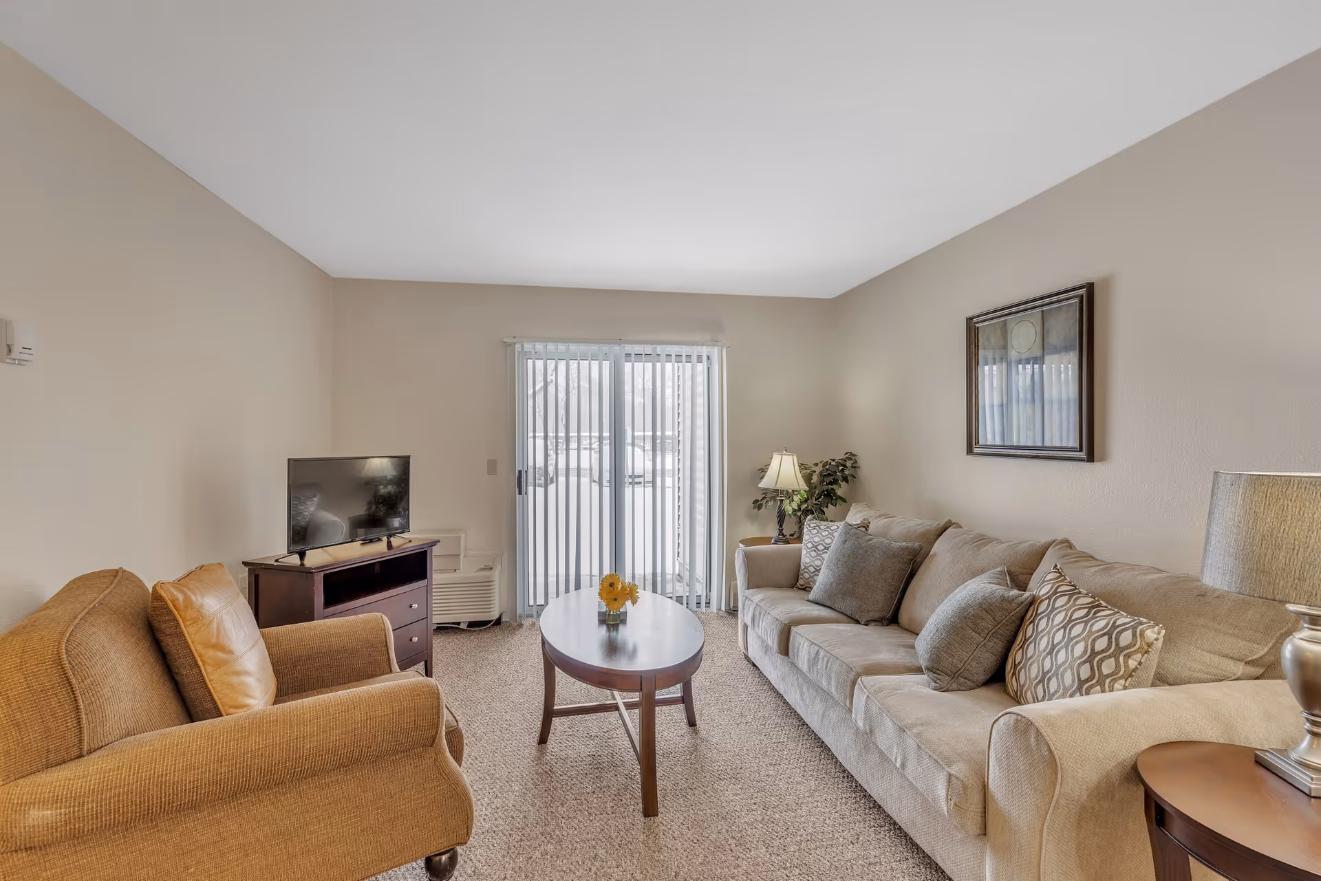 Cozy living room with a beige sofa, armchair, coffee table, TV on a stand, and sliding glass doors with vertical blinds.