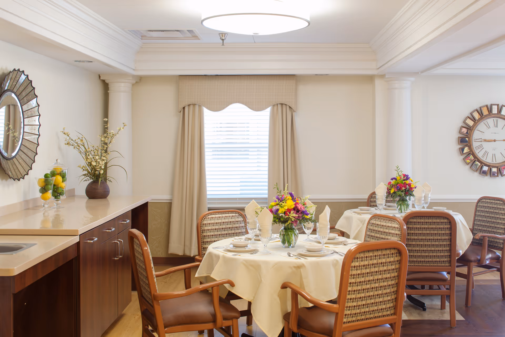 A bright dining room with round tables covered in white tablecloths, each set with plates, glasses, napkins, and floral centerpieces. The room features wooden chairs with patterned upholstery, a large window with beige curtains, decorative wall mirrors, and a countertop with a vase of flowers and a glass container filled with lemons and limes.