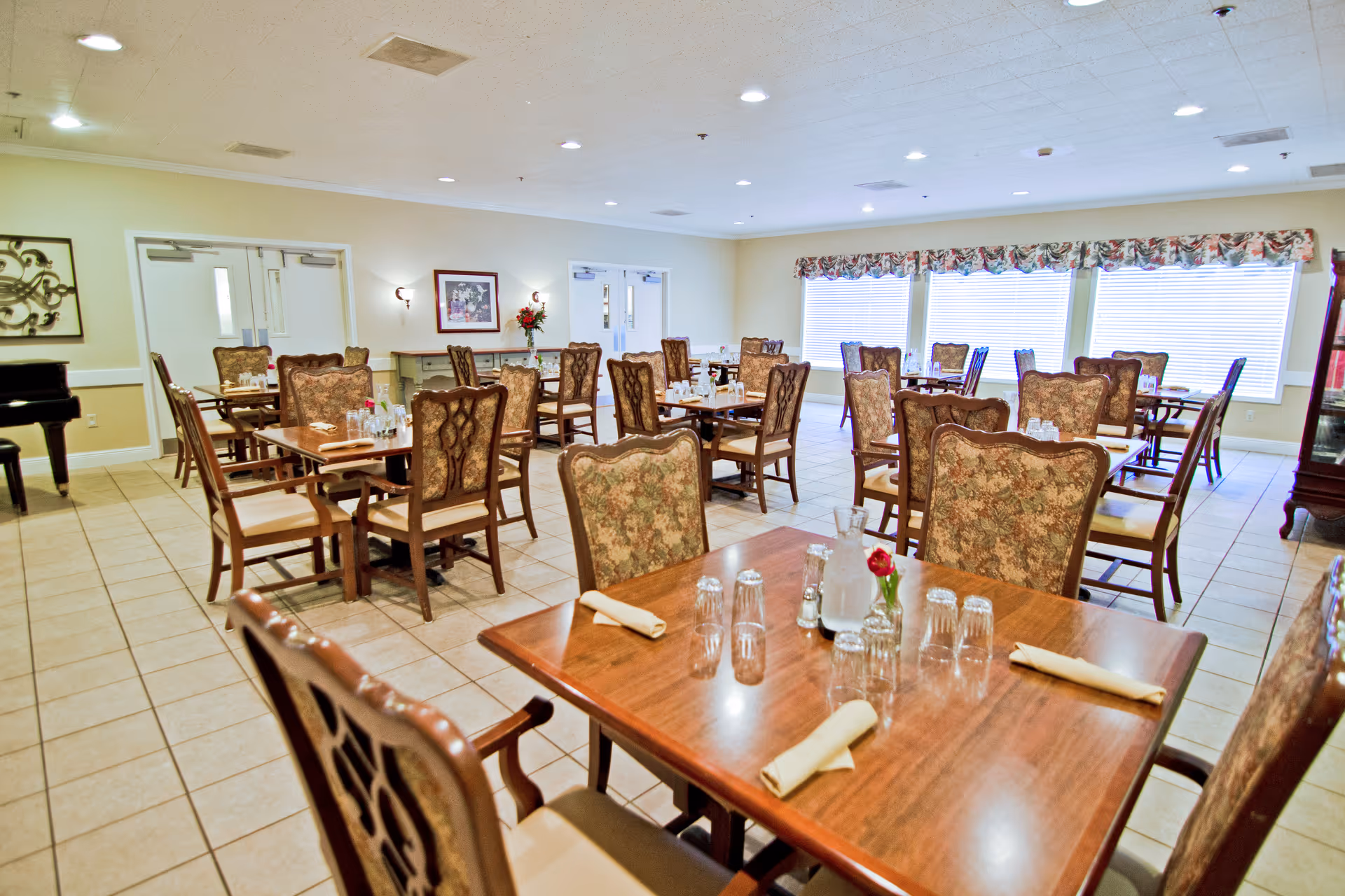 A bright dining room with multiple wooden tables and upholstered chairs arranged neatly. Each table is set with glasses, napkins, and small flower vases. Large windows with floral valances allow natural light to fill the room. The floor is tiled, and the walls are painted a light color. There is a piano visible in the corner and framed artwork on the walls.