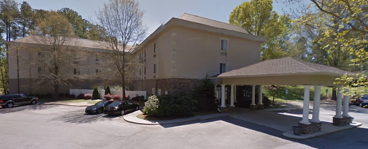 Front exterior of a multi-story senior living building with a covered porte-cochere, parked cars, and surrounding trees.