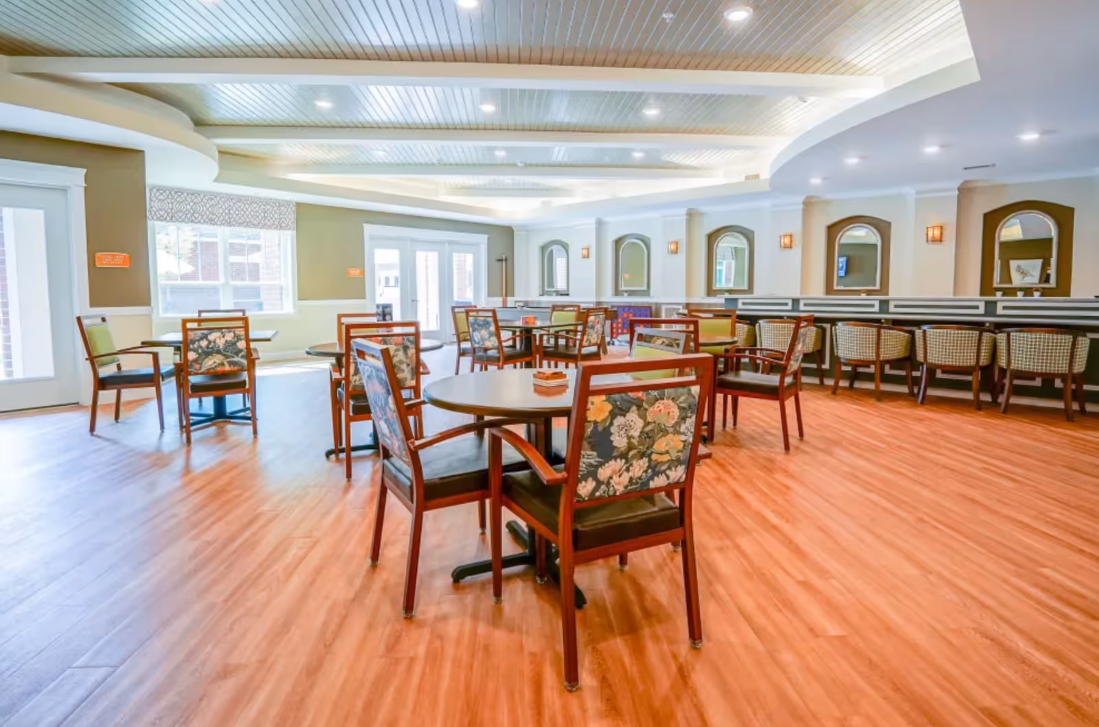 Bright dining room in a senior living facility with round tables, floral-upholstered chairs, and bar seating.