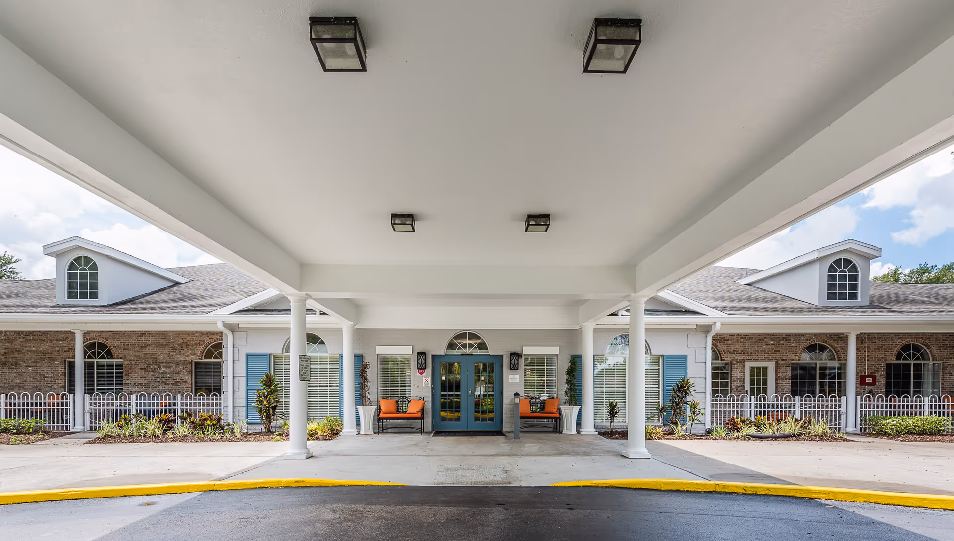 Covered entrance area of a senior living facility with white columns supporting the roof. Two benches with orange cushions are placed on either side of a blue double door entrance. The building has brick and white walls with blue shutters and arched windows. There is a small garden with plants and a white fence in front of the building.