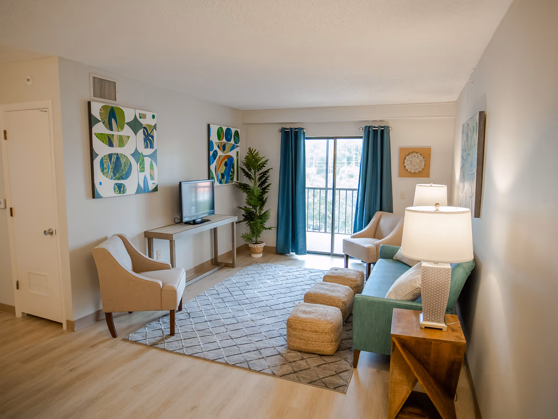 A bright and cozy living room in an assisted living community featuring a teal sofa with a white pillow, two beige armchairs, three woven ottomans, a gray patterned rug, a wooden side table with a lamp, a console table with a flat-screen TV, two abstract paintings on the walls, a potted plant, and large sliding glass doors with teal curtains leading to a balcony.