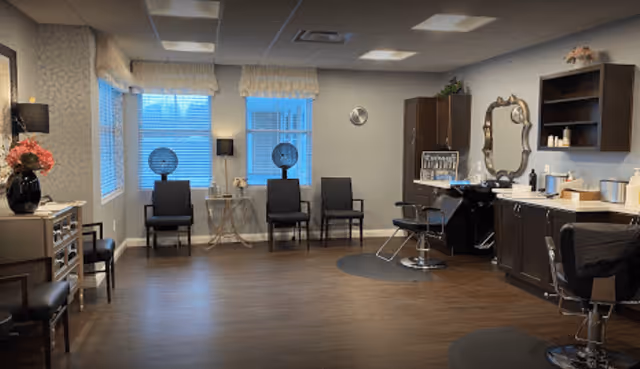 A spacious salon area with wooden flooring, featuring multiple black salon chairs, hair drying stations with hooded dryers, a large ornate mirror, and cabinets with various hair care products. The room has large windows with blinds and soft lighting from ceiling fixtures and lamps.