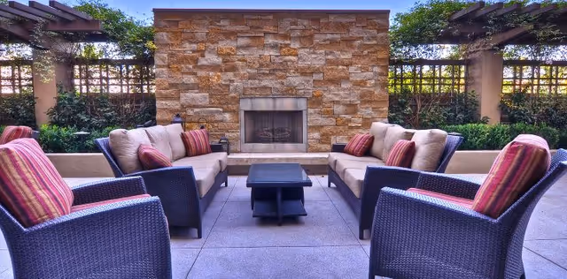 Outdoor seating area with cushioned wicker chairs and sofas arranged around a central black coffee table in front of a stone fireplace, surrounded by greenery and lattice fencing under a pergola.