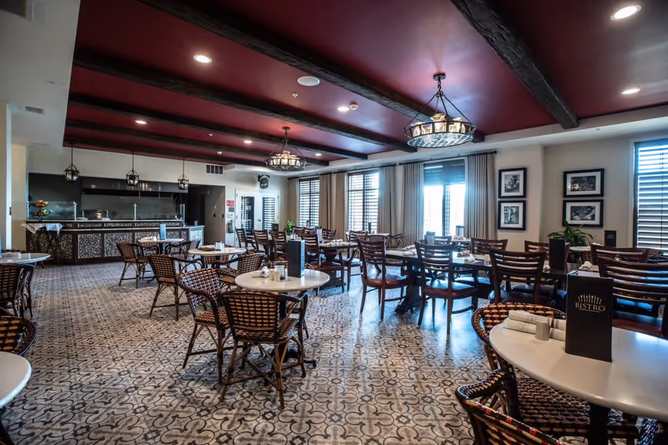 A spacious dining room with multiple round and rectangular tables surrounded by wooden chairs. The room features patterned tile flooring, a dark red ceiling with exposed wooden beams, and large windows with curtains allowing natural light. A counter area is visible at the back with hanging pendant lights and decorative elements.