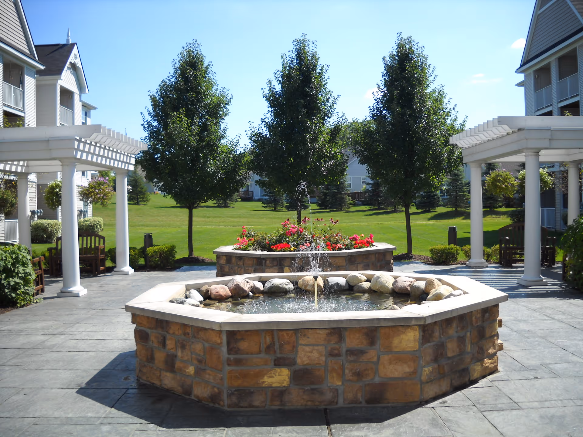 Outdoor courtyard area at Oakhaven Manor featuring a stone octagonal water fountain with a small water jet, surrounded by a paved patio. There are two white pergolas with benches underneath on either side of the fountain. In the background, there are green trees, a well-maintained lawn, and multi-story residential buildings.