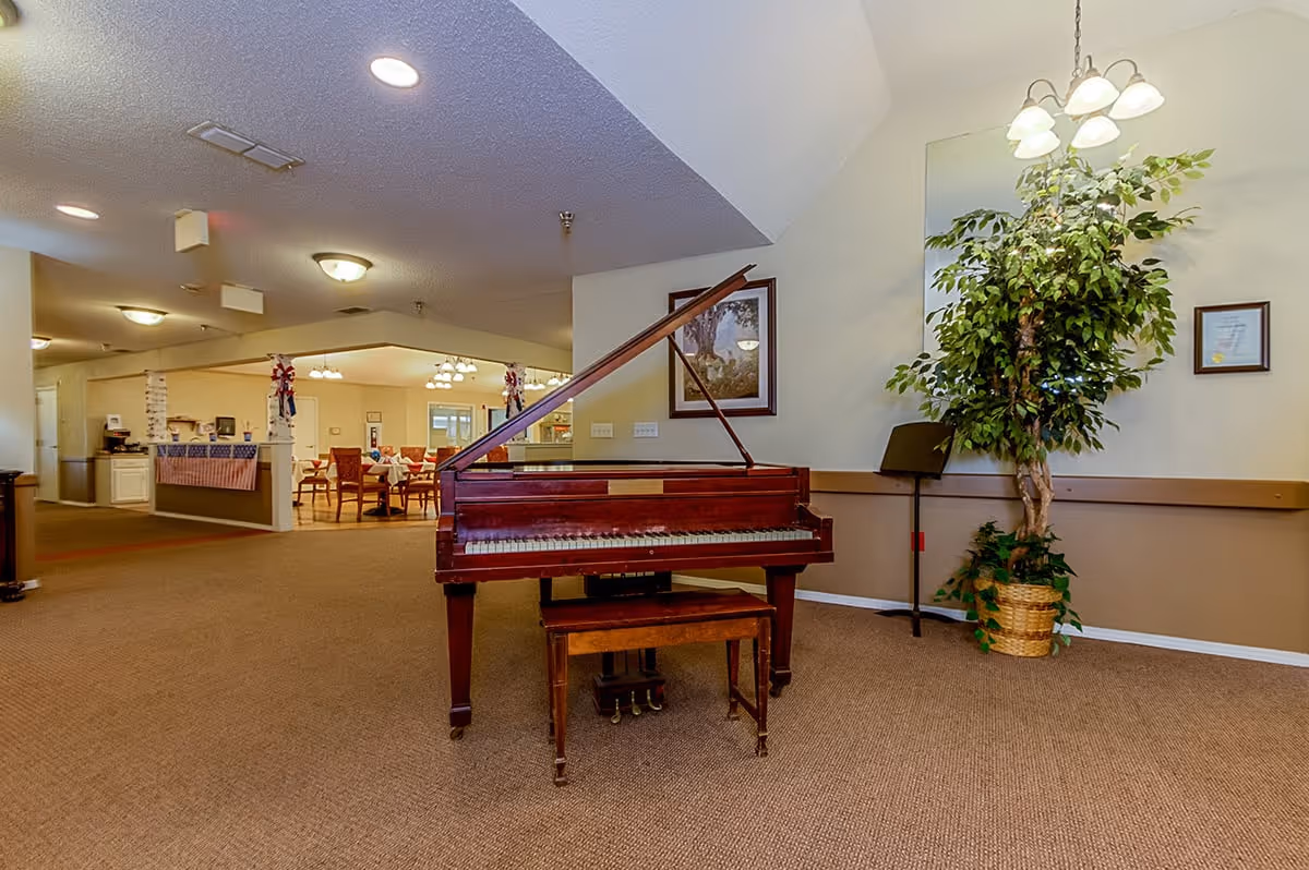 Indoor common area of a senior living facility with a grand piano and bench, a potted plant, and a dining area visible in the background.