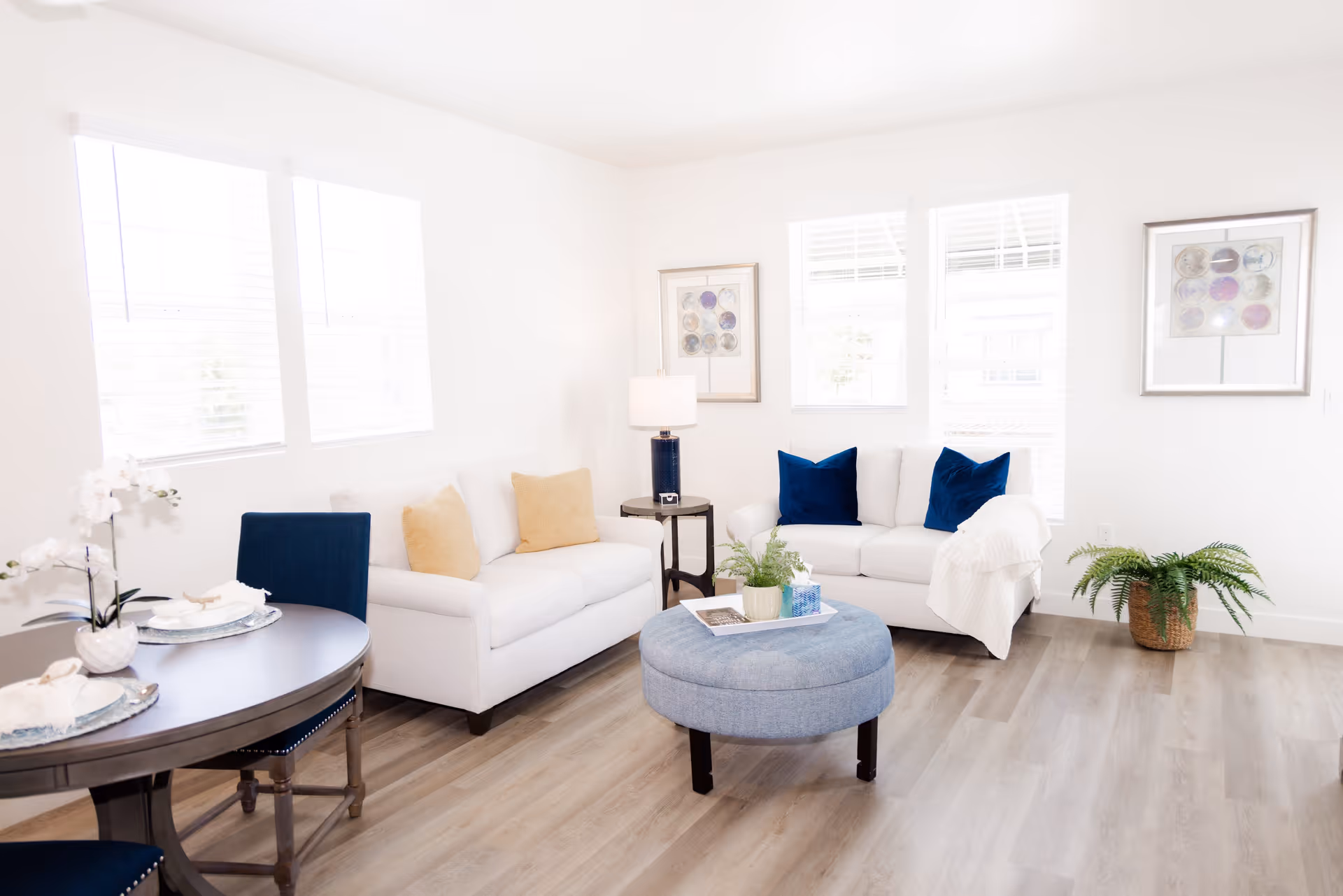 Bright living room with two white sofas adorned with blue and beige pillows, a round blue ottoman in the center, a small side table with a lamp, two framed artworks on the wall, large windows letting in natural light, a round dining table with blue chairs, and a potted plant on the floor.