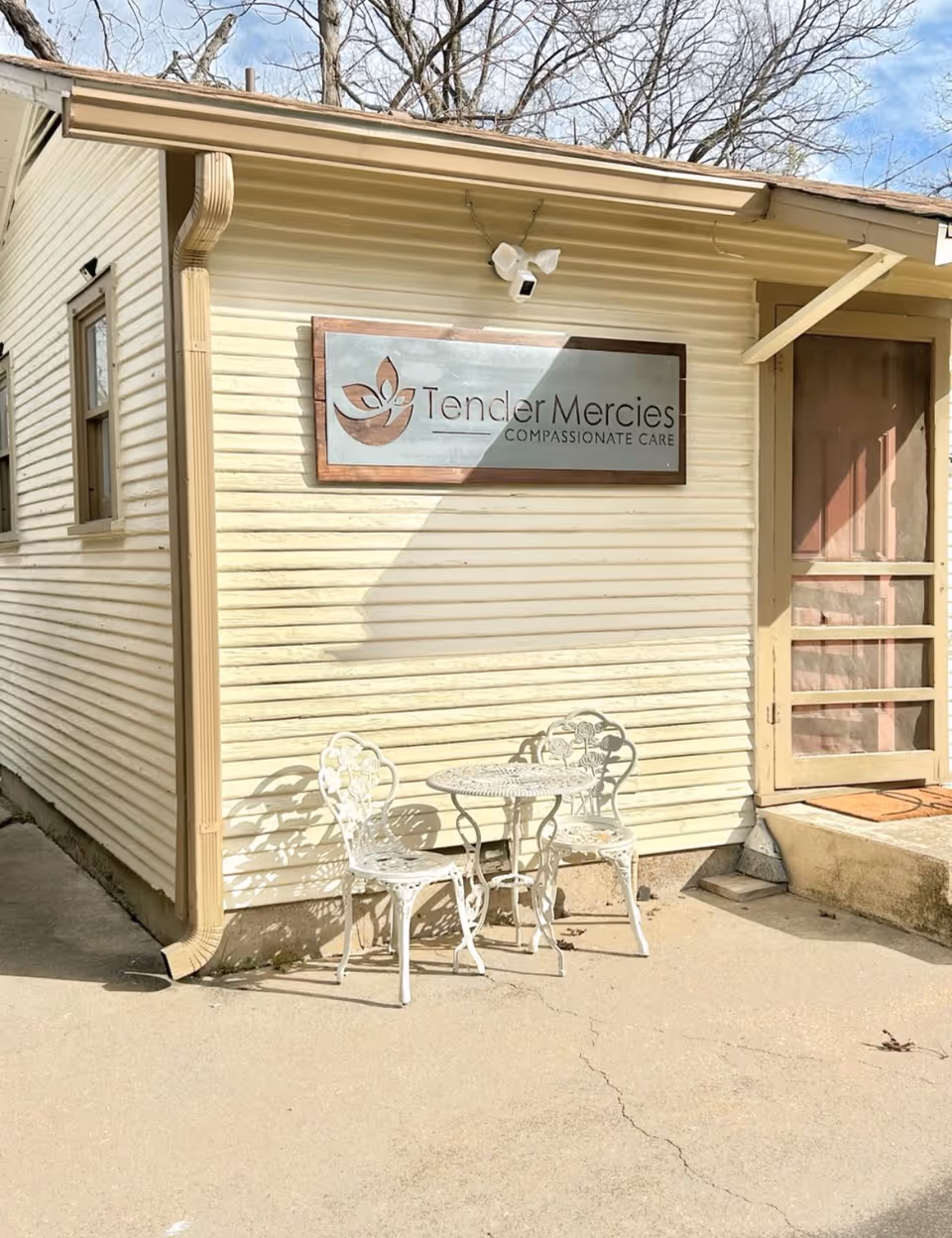 Exterior view of a light yellow building with a sign that reads 'Tender Mercies Compassionate Care'. In front of the building, there is a small white metal table with two matching chairs on a concrete surface. Leafless trees are visible in the background under a partly cloudy sky.