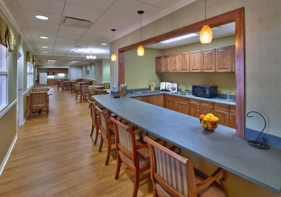 A spacious dining area with wooden floors and multiple tables and chairs arranged along the left side. On the right side, there is a kitchen counter with several chairs, pendant lights hanging above, wooden cabinets, a microwave, and a bowl of fruit on the counter. The walls are painted green and the ceiling has recessed lighting.