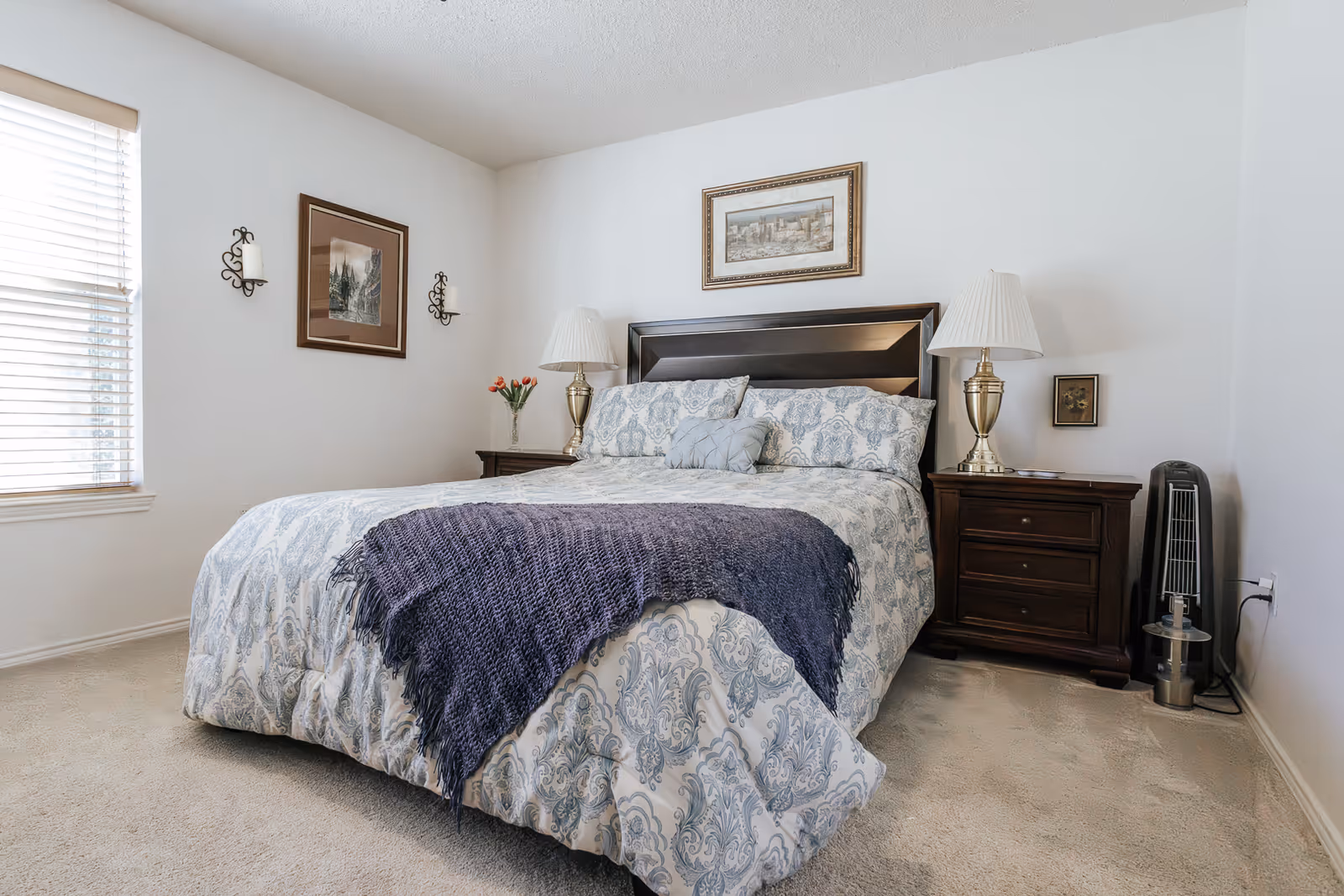 A neatly made bed with a patterned comforter and a dark purple throw blanket in a bedroom. The bed has a dark wooden headboard and is flanked by two matching nightstands, each with a lamp. There are framed pictures on the white walls and a window with blinds letting in natural light.