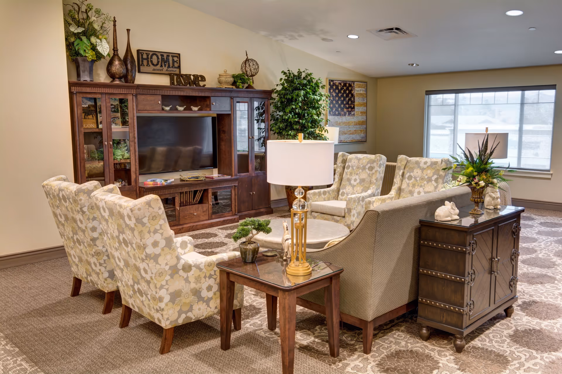A cozy living room area in an assisted living facility featuring a large wooden entertainment center with a flat-screen TV, decorative items including a 'HOME sweet home' sign and 'LOVE' letters, floral patterned armchairs, a beige sofa, side tables with lamps and small plants, a large window with blinds, and a framed American flag artwork on the wall.