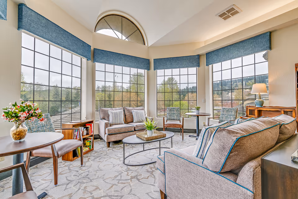 Bright and spacious living room area with large windows offering a scenic view of trees and hills outside. The room features comfortable beige sofas with blue piping, a round coffee table with a plant centerpiece, a small bookshelf, and several chairs around small tables. Blue window valances complement the neutral tones of the room, and a table lamp adds warmth to the cozy setting.