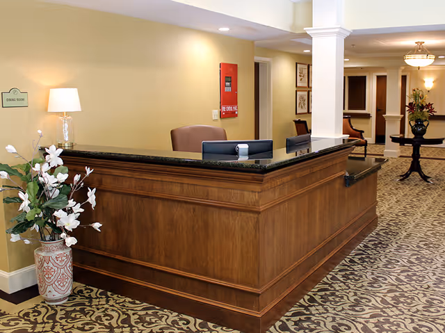 Reception desk area with a wooden counter and black granite top, two computer monitors, a brown chair, a table lamp, a large floral arrangement in a decorative vase, and a sign on the wall indicating the dining room. The background shows a hallway with framed artwork, chairs, a round table with a flower arrangement, and warm lighting.