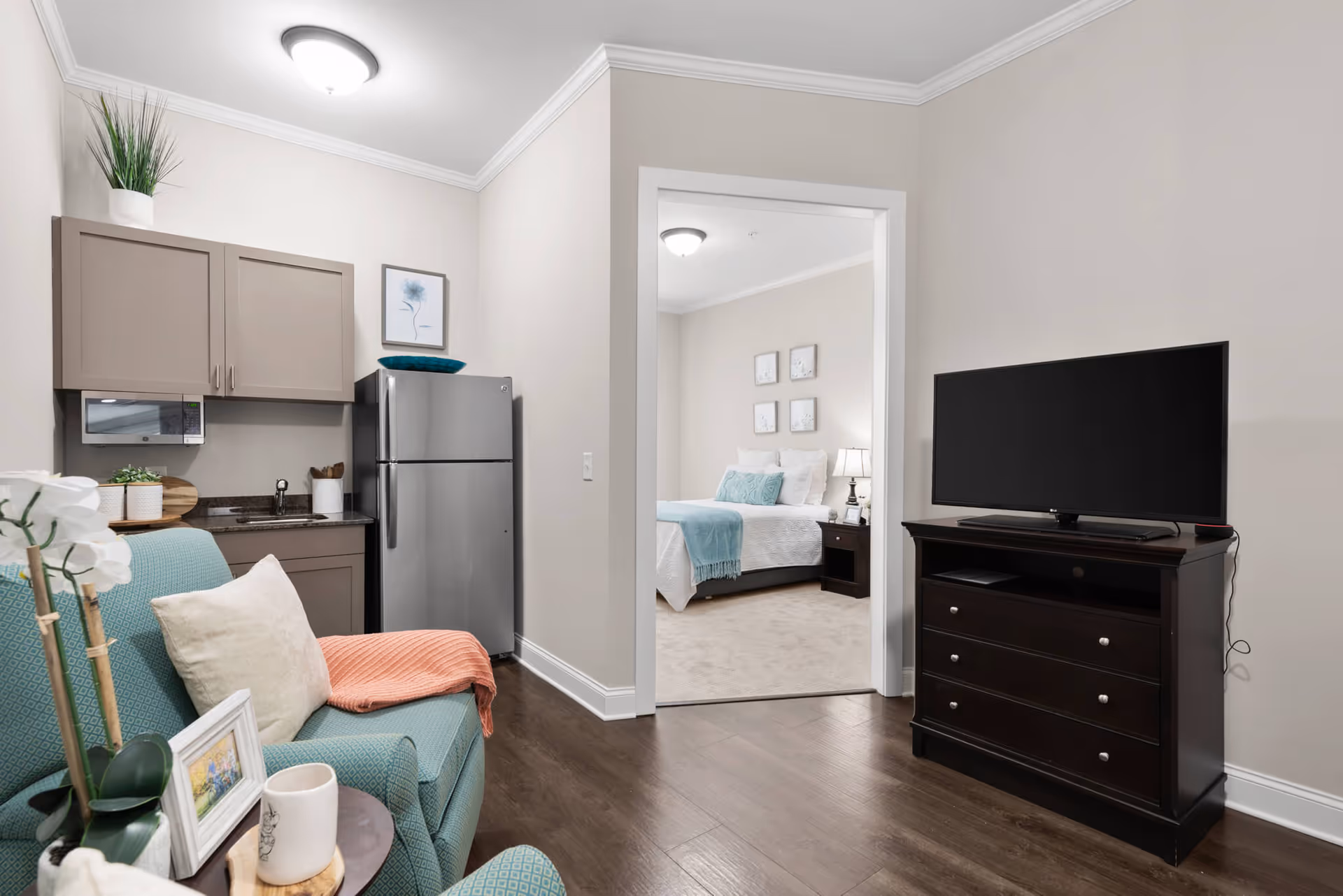 A cozy living area in a senior living facility featuring a small kitchenette with a refrigerator, microwave, and cabinets. There is a teal armchair with a white pillow and a coral throw blanket. A flat-screen TV is placed on a dark wooden dresser. Through an open doorway, a bedroom with a neatly made bed, nightstand, and lamp is visible.